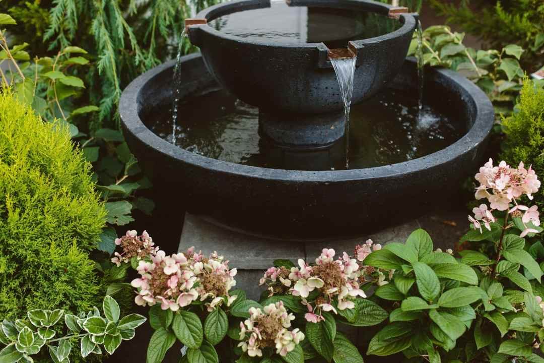 Black tiered round fountain surrounded by plants and hydrangeas - 7 Dees