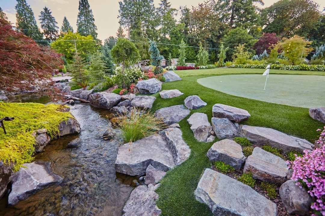 Naturalistic manmade stream meanders past boulders and a putting green with trees in the background 7 Dees Landscaping